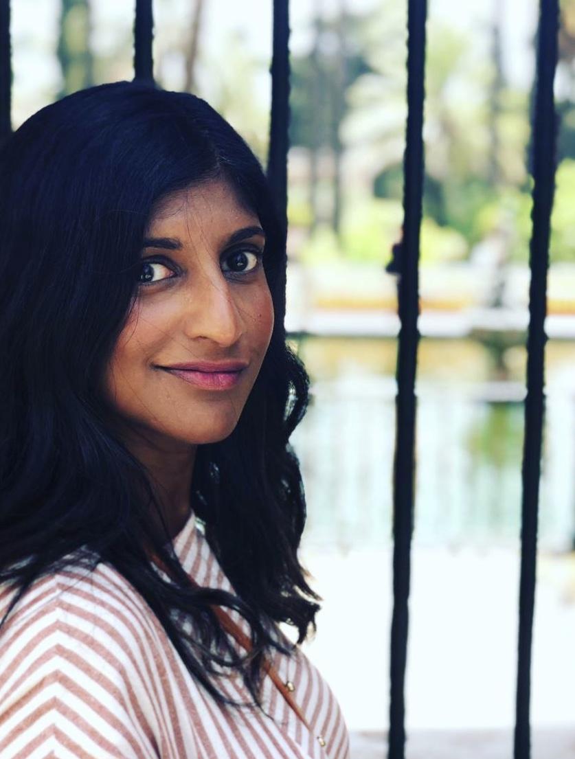 Therapist Ancy Cherian looking over her shoulder smiling at the camera wearing a pink and white striped shirt