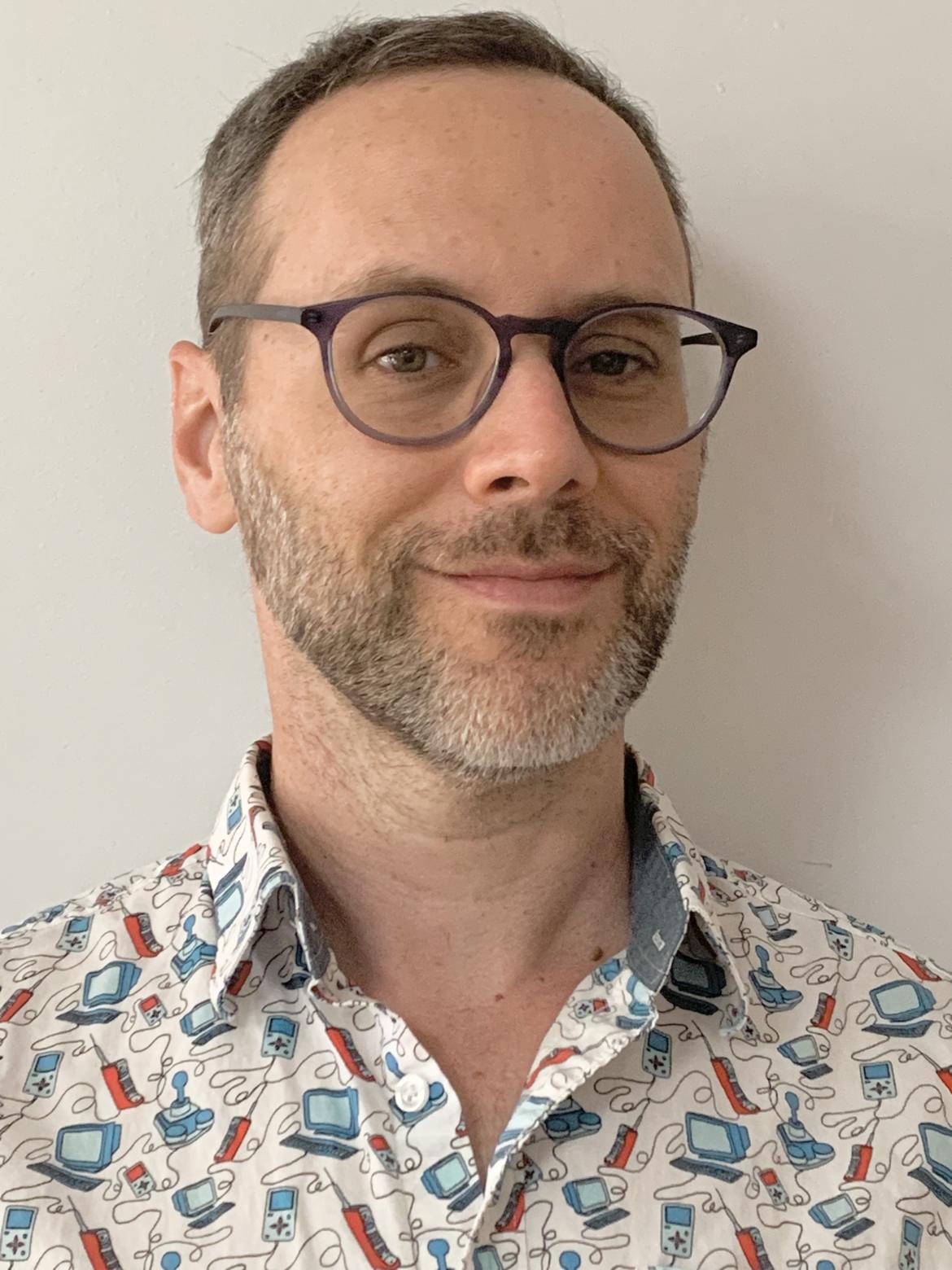 Therapist Charlie Padow smiling at the camera wearing glasses and a white button up shirt with little computers and retro controllers