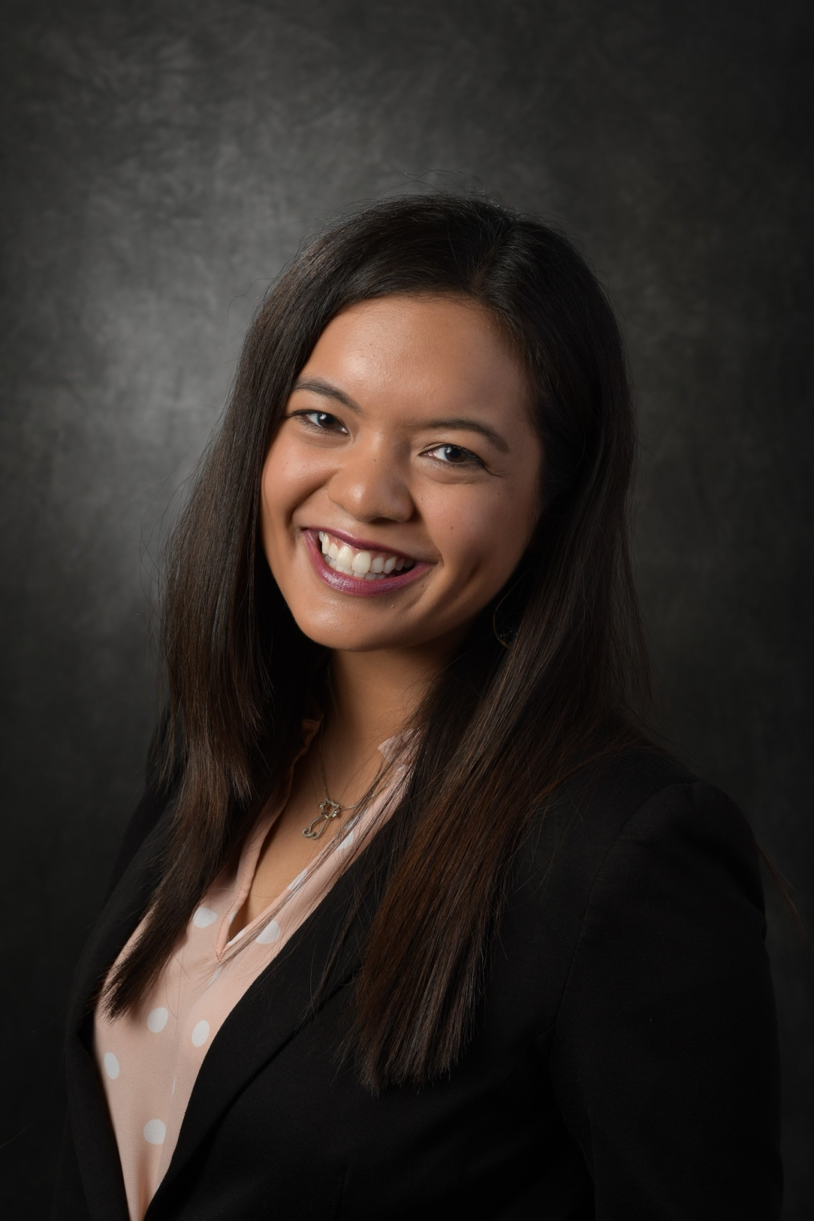 Therapist Catherine Teotico wearing a pink shirt with white polka dots and a black blazer with a dark grey background, smiling at the camera