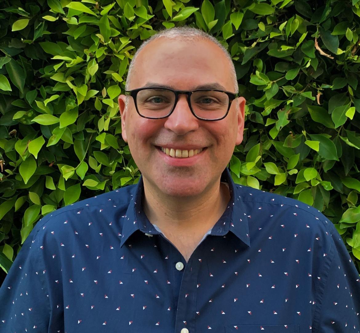 Therapist Jason Tomasian wearing a blue shirt with while dots on it smiling at the camera in front of a wall of leaves