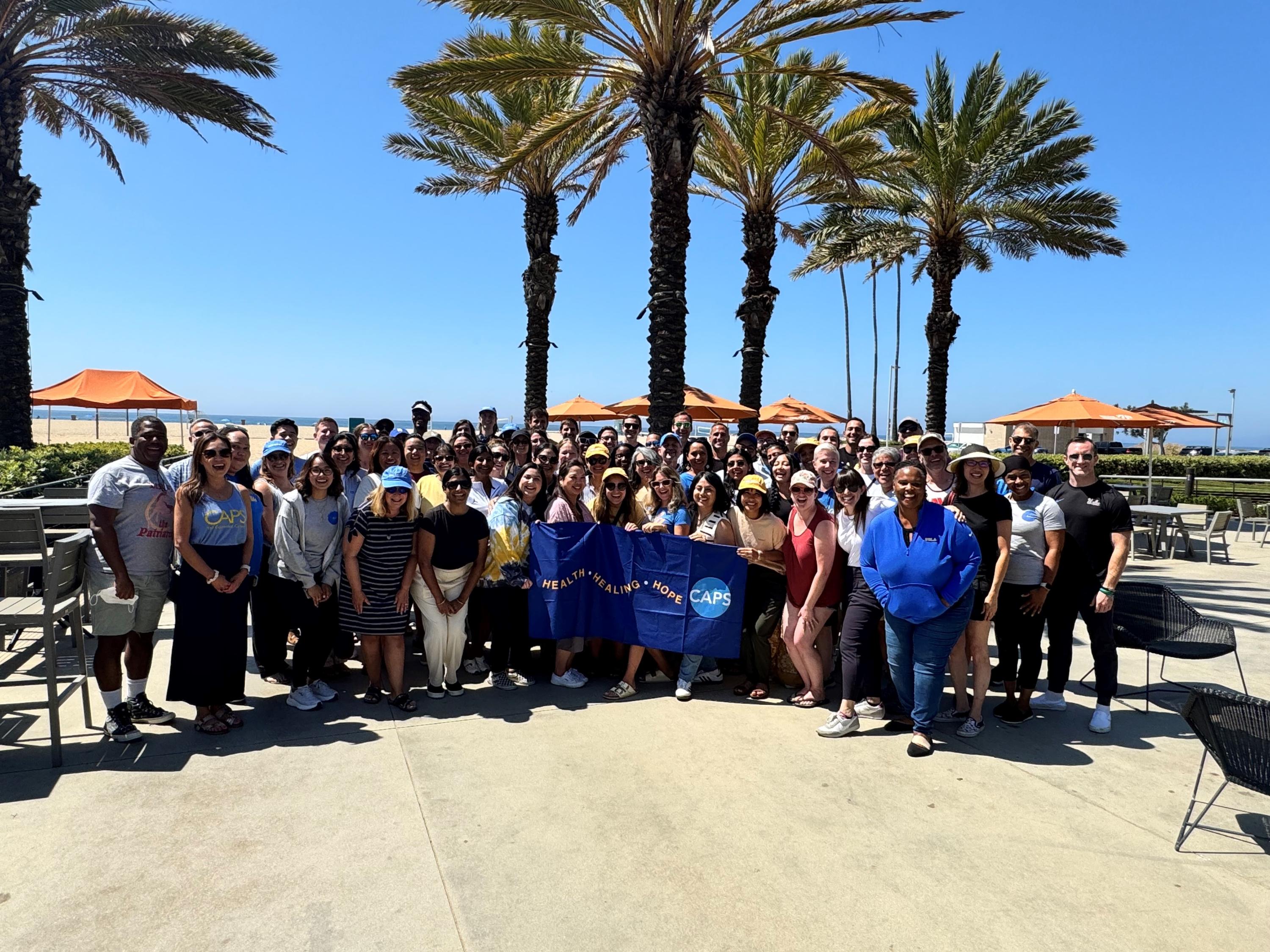 CAPS staff smiling at the camera at their annual staff retreat in Santa Monica. The beach is behind them and they are holding a towel that reads heath, healing, hope.