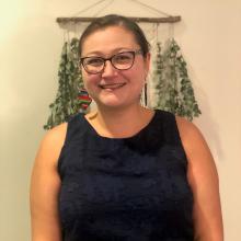 Therapist Juliana Carranza wearing a blue tank top smiling at the camera with green leaves hanging on the wall behind her