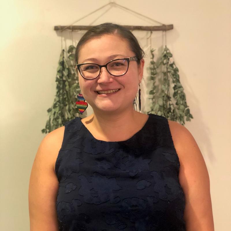Therapist Juliana Carranza wearing a blue tank top smiling at the camera with green leaves hanging on the wall behind her